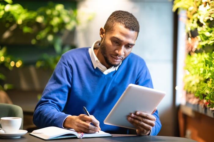 A man sitting in a cafe surrounded by green plants and writing in a notebook while holding up a tablet.